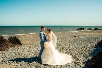 Bride and groom together on a pebble beach with the sea and a rocky headland behind them