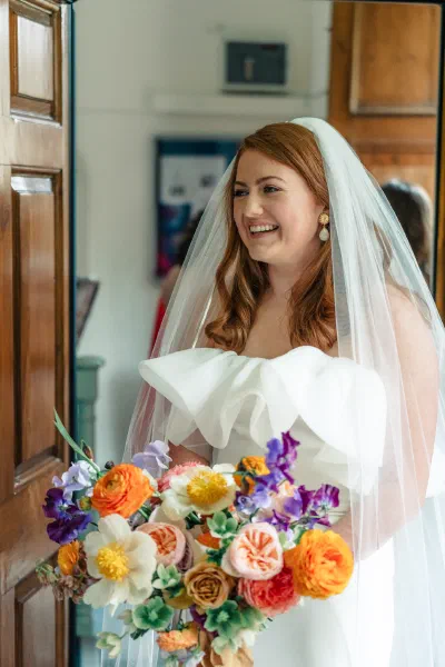 Bride laughing in the church doorway in a strapless dress with a large folded bodice detail, holding a bright spring bouquet