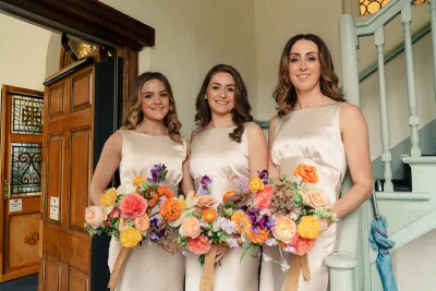 Three bridesmaids in oyster satin dresses holding bouquets of orange, coral and purple flowers in a doorway