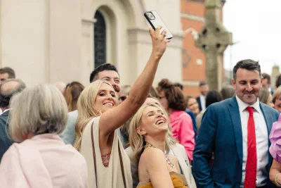 Wedding guests outside the church, one holding a phone in the air, laughing after the ceremony