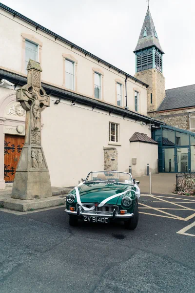 Vintage green MG convertible with white ribbon parked beside a carved Celtic high cross outside the Church of the Assumption in Booterstown