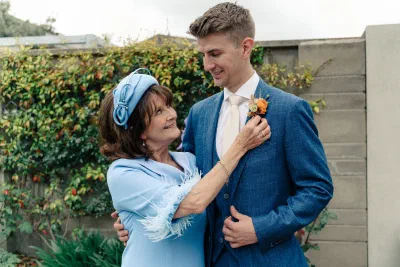 Groom’s mother in a pale blue dress and headpiece pinning an orange buttonhole to his blue check suit in a Dublin garden