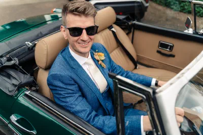Groom in a blue check suit and sunglasses sitting behind the wheel of a vintage green MG convertible outside a house in Dublin