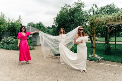 Bride laughing as two friends hold her long veil out behind her on a gravel path in the walled garden