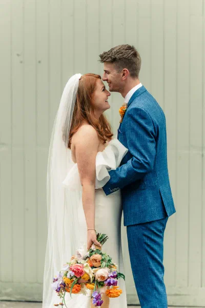 Bride and groom kissing against a wall of pale sage painted timber boards, her bouquet at her side