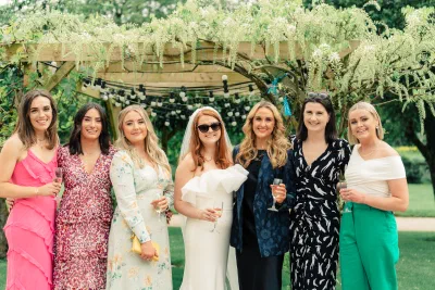 Wedding guests in bright pink, green and floral outfits standing under a wisteria covered pergola in the garden at The Millhouse