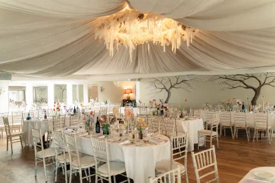 The orangery at The Millhouse set for a wedding dinner, with a draped ceiling, chandeliers and a hand painted tree mural along the wall