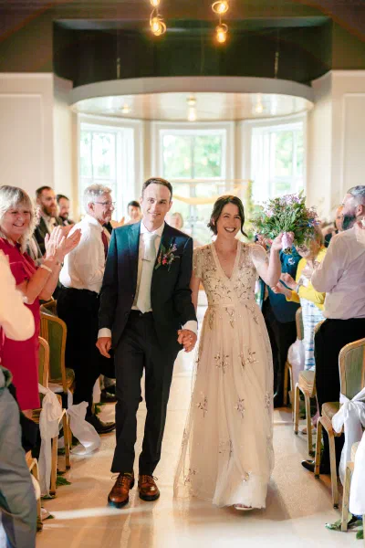 Newlyweds walking back up the aisle hand in hand as guests applaud, Orlagh House ceremony room