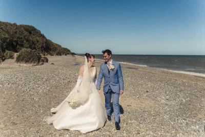 Wide photograph of the bride and groom walking a deserted beach beneath a headland