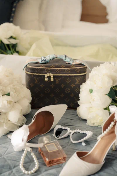 Bridal shoes, a pearl bracelet, Chanel perfume, heart shaped pearl sunglasses and white peonies arranged on a bed beside a monogrammed vanity case