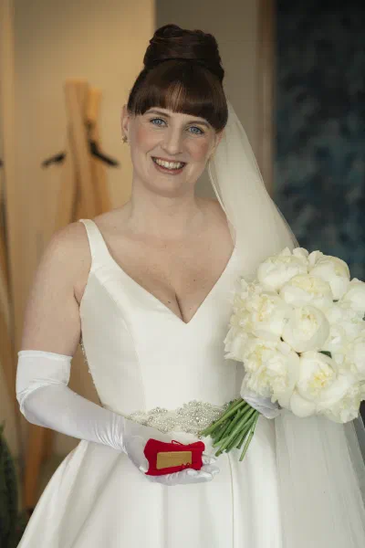 Bride seated on the end of a four poster bed in her satin wedding dress, holding a white peony bouquet