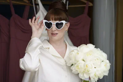 Bride in cream satin pyjamas wearing heart shaped pearl sunglasses and holding a white peony bouquet