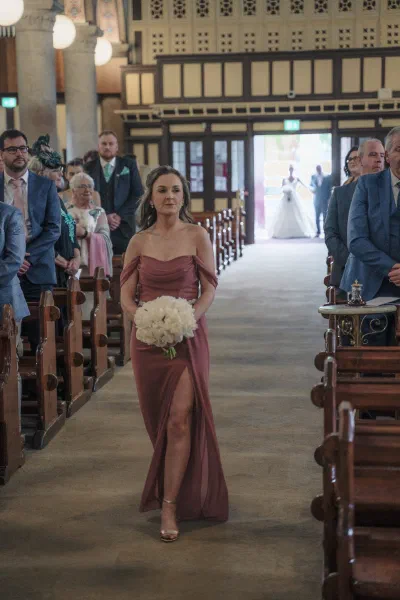 Bridesmaid in a dusty rose dress walking down the aisle carrying white peonies