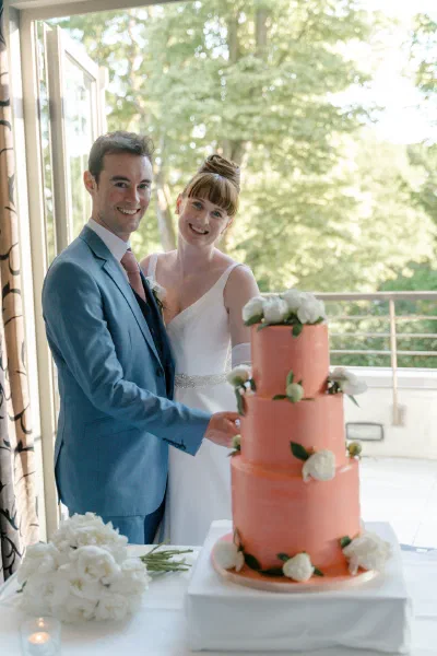 Bride and groom cutting their coral pink wedding cake in front of a tall window