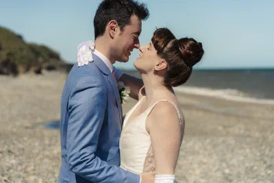 Bride and groom about to kiss on a pebble beach, a green headland and the Irish Sea behind them