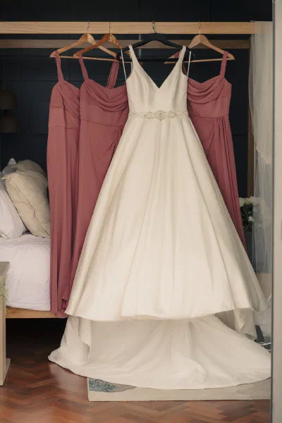 A white wedding dress hanging alongside four dusty rose bridesmaid dresses in a hotel bedroom at Seafield