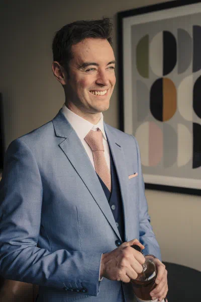 Groom in a cornflower blue three piece suit with a dusty pink tie, standing by a window