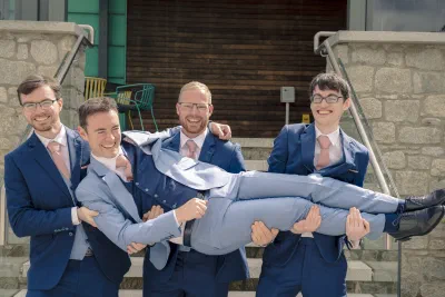 Four groomsmen in navy suits lifting the groom horizontally outside the hotel, everyone laughing