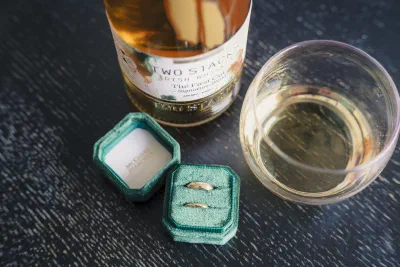 Two gold wedding bands in a green velvet ring box beside a bottle of Two Stacks Irish whiskey and a filled glass