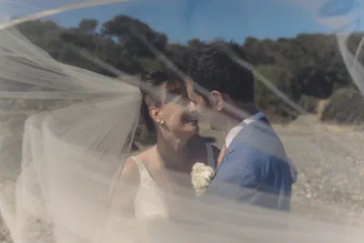 Bride’s veil blown across both of them by the wind, the couple looking at each other underneath it