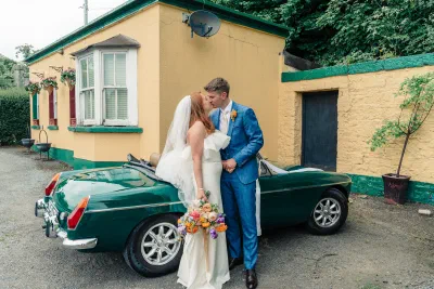Bride and groom leaning against the vintage green MG outside a yellow painted cottage with green trim